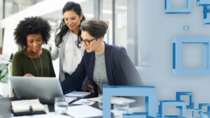 A group of business women look at something on a laptop screen in an office.