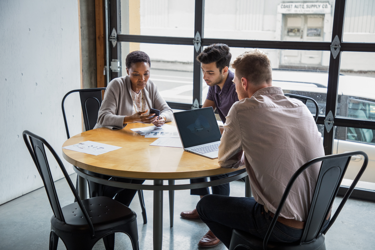 One female and two male workers sitting at round table in casual office setting or café/coffeehouse. The woman is holding a phone to show something to one of the men. The other man is using a Surface Laptop.