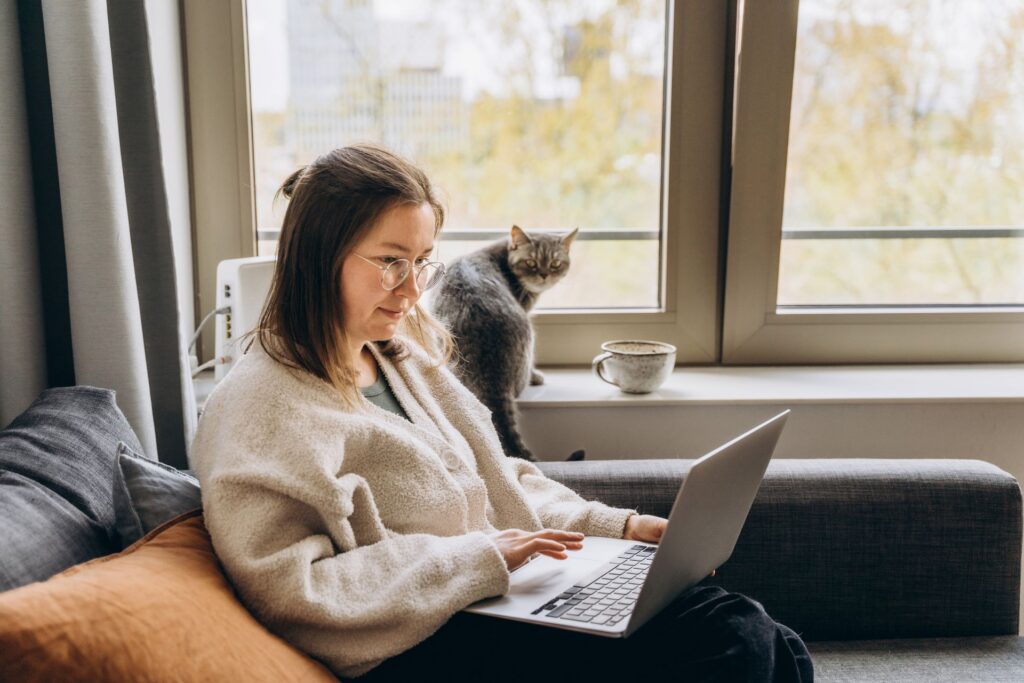 An educator sits on a sofa while looking at a laptop screen. There’s a cat nearby.