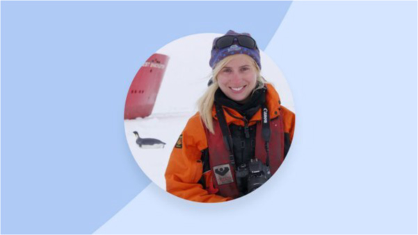 A woman with blonde hair wearing a knit hat, orange parka, and a camera stands on a patch of ice with a penguin and ship in the background.