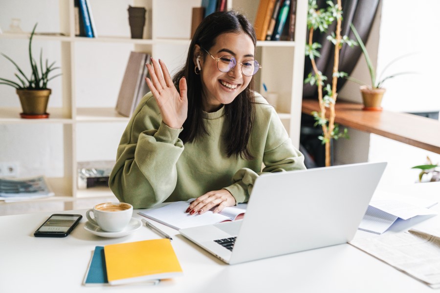A college student is engaged in a video call and waving at the camera from a desk in shared workspace.