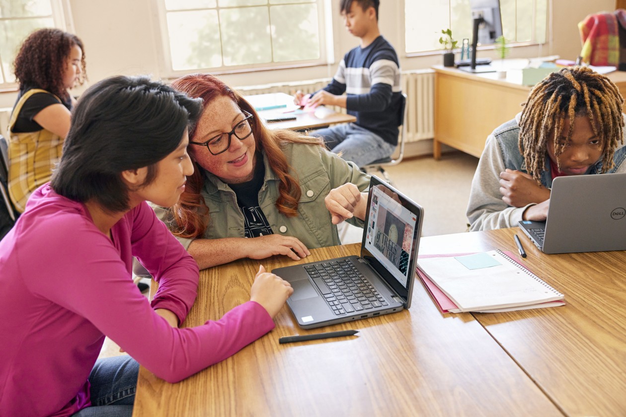A classroom scene with students at communal desks and an educator leans in to help one of the students.