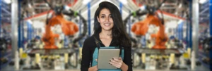 A person looking at the camera smiling and holding a tablet. There is manufacturing machinery in the background.