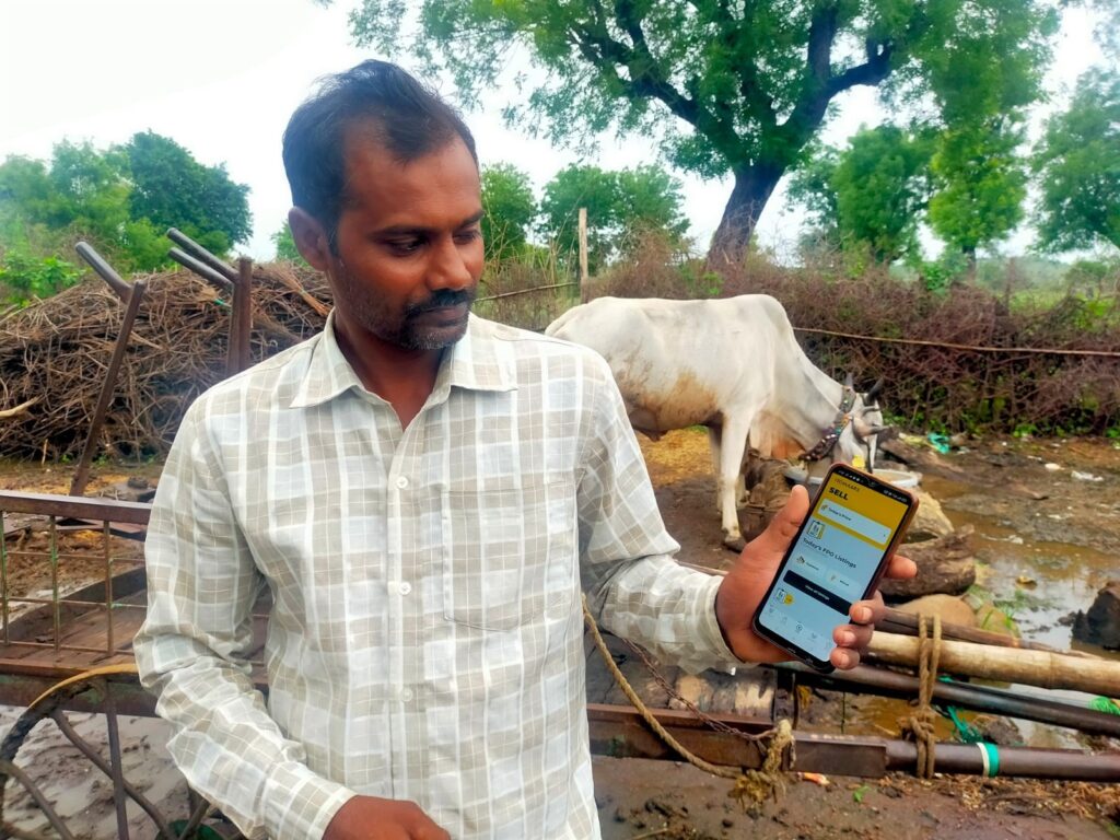 Image of a person holding a mobile phone with the ITC Krishi Mitra app. In the background, there is a plow, a cow is drinking water, and several trees.