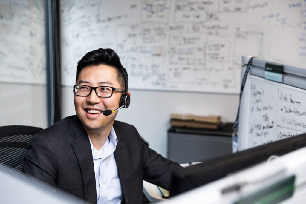 Male worker sitting at cubicle desk in financial office, wearing glasses and phone headset. He is smiling and looking up to his right. A large whiteboard covered in black written text is on wall behind him. A smaller whiteboard also partially visible on his desk's cubicle wall.