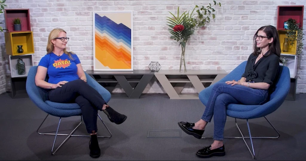 Two women, Di Mayze and Kate Rosenshine sit facing each other in armchairs. There are bookshelves, plants, and paintings behind them.