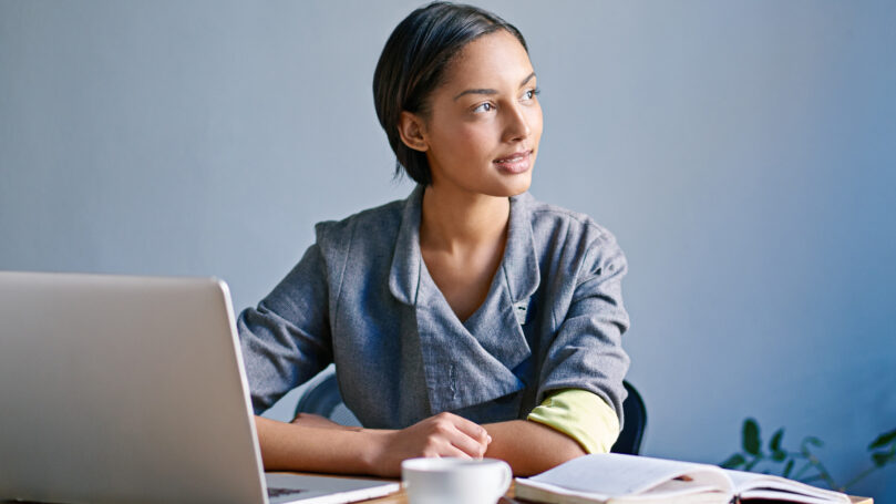 A woman is sitting at a wooden table with a laptop, an open book, and a cup of coffee. The background is plain and the person is wearing a grey jacket.