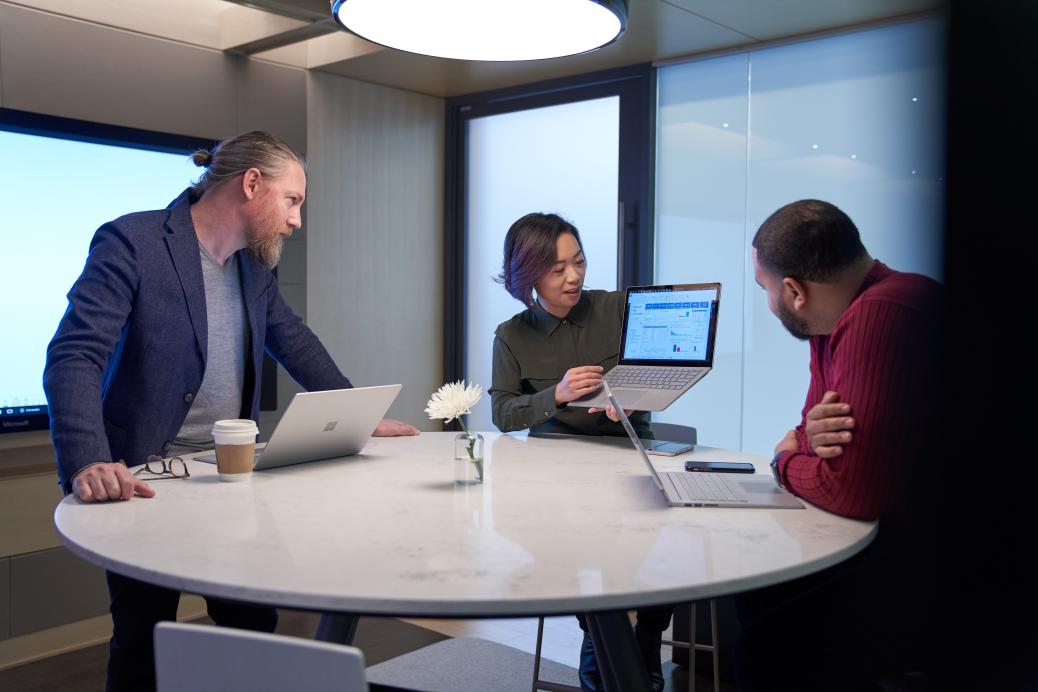 Woman and two men working in a meeting room all with Surface laptops.