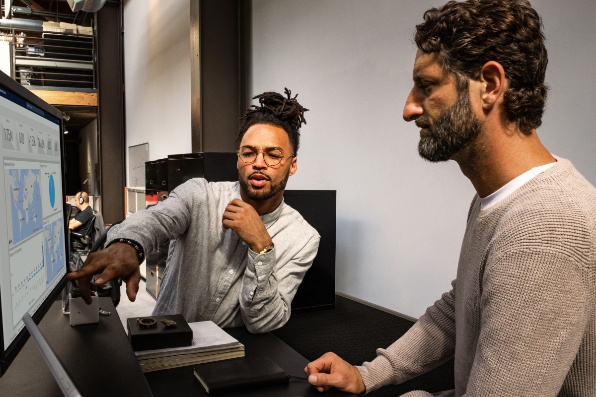 Two adult males working at computer station together.