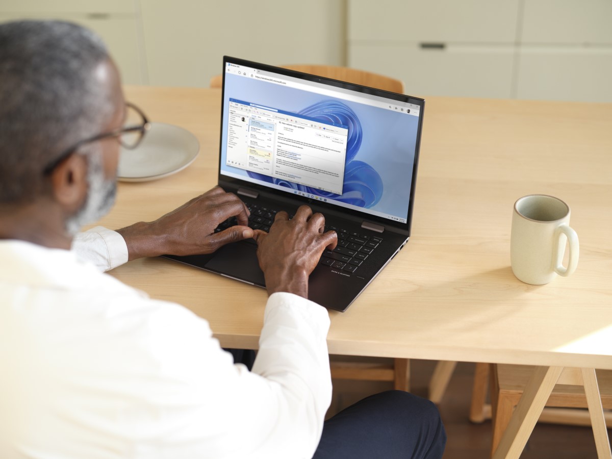 Man sitting at a wooden table typing on a laptop while using Microsoft Teams within Windows 365. Shown on Windows 11.