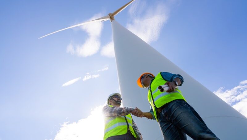 Two field workers shake hands beneath a tall wind turbine