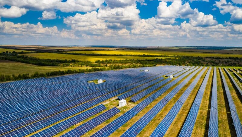 An expansive solar array stretches across fields, under a bright sky.