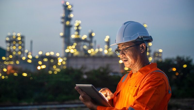 An engineer wearing a hardhat uses a digital tablet while working the night shift at petroleum oil refinery.
