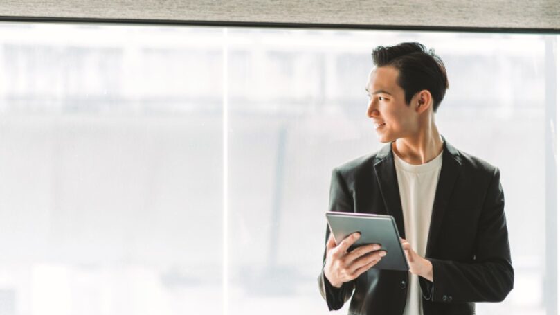 An young businessman holding a digital tablet stands in front of a large window in his modern office.