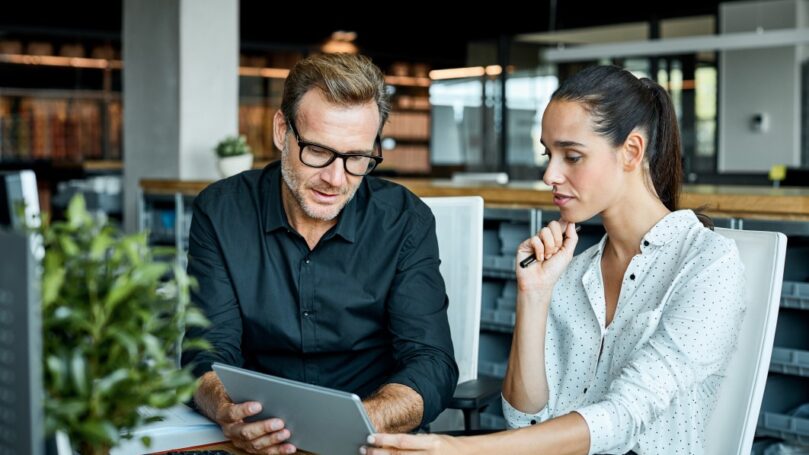 A businesswoman reviews a colleague's presentation on a digital tablet at her desk in an open office space.