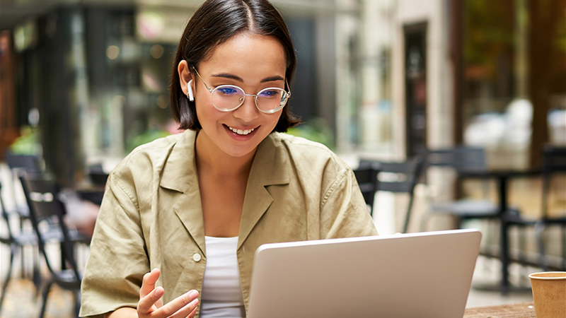 Una mujer con gafas y auriculares sonríe mientras usa una computadora portátil al aire libre.