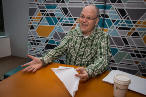 Apple sits at a table while holding a paper airplane and talks to someone off-screen