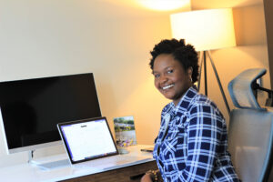 Nyameke sits in front of her laptop and monitor while smiling at the camera.