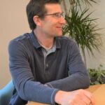 Martin sits at a table and looks to the left as he talks to someone.