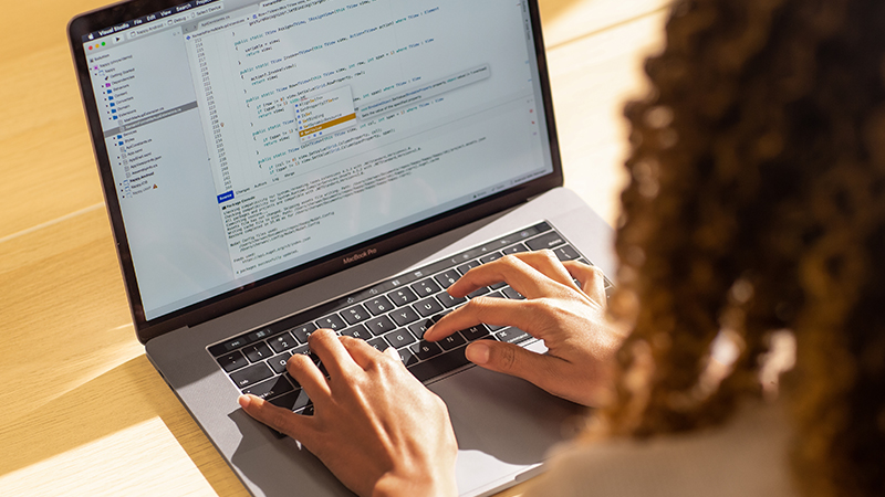 Pessoas reais, escritórios reais. Desenvolvedora negra codificando em um MacBook Pro no escritório, usando Visual Studio.
